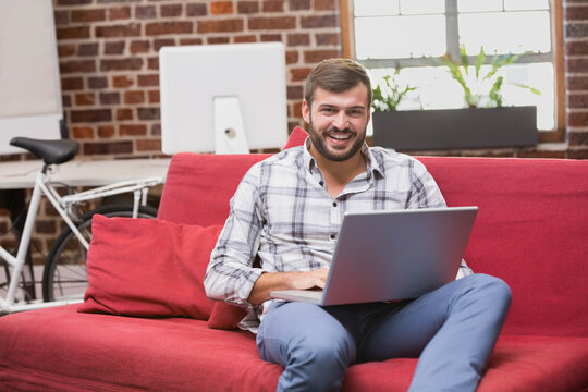 Male professional smiling toward camera while working on laptop on red sofa in loft home office - Powered by Adobe