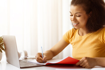 Smiling African American woman writing notes in red notebook at laptop desk. Remote learning, freelance projects or modern online student lifestyle at home