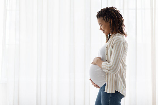 Side view of smiling pregnant Black woman touching belly, standing near bright curtains at home, enjoying maternity anticipation with peaceful lifestyle tone