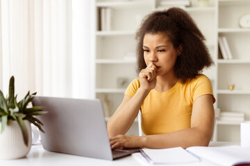 Serious African American woman concentrating while working on laptop at home office desk. Concept of remote work challenges, online studying or business planning