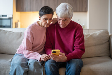 Concerned elderly couple checking smartphone on couch. Seniors read suspicious message, husband wife phone scam victim, online fraud, stress from message, risk digital security concerns for seniors