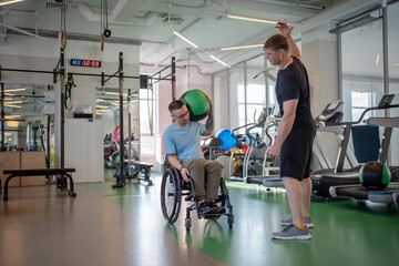 Serious focused disabled man do ball lifts under coach guidance in gym. Rebuilding neuromuscular connections after traumatic paralysis, post trauma athletic recovery, neurological recovery training