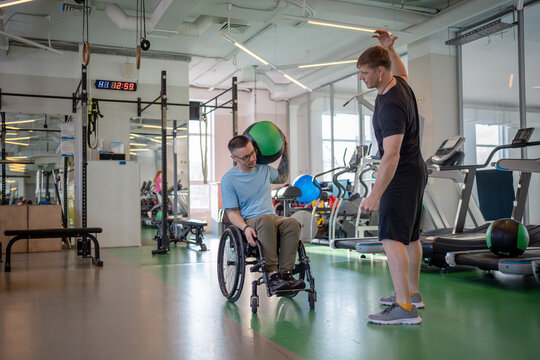 Serious focused disabled man do ball lifts under coach guidance in gym. Rebuilding neuromuscular connections after traumatic paralysis, post trauma athletic recovery, neurological recovery training