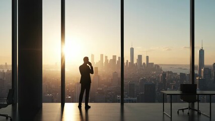 Silhouette of a businessman standing by a large office window, talking on his cell phone while looking at the city skyline during a beautiful sunrise or sunset - Powered by Adobe