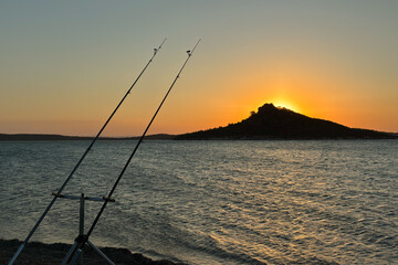 Peaceful sea fishing with a rod against clear blue sky or sunset  and calm ocean water, tranquil coastal landscape with distant mountains, summer outdoor leisure and recreational activity concept.