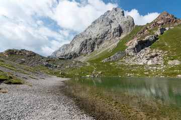 Rocky shoreline of Volaia lake (Wolayersee) with crystal-clear waters reflecting the surrounding peaks, under a bright summer sky in the pristine alpine landscape of the Italy-Austria border.