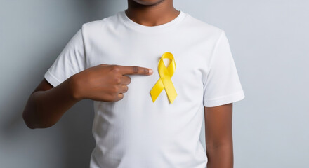 African American boy stands with a yellow ribbon on a white t-shirt, pointing at it. Design template for childhood cancer awareness month concept and support for disease patients. 