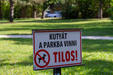 Close-up view of Hungarian-language park sign forbidding dog walking on grass, with blurred sunlit lawn in background. Budapest, Hungary