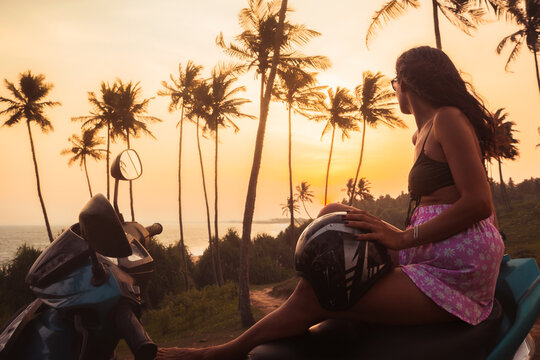 Carefree pretty woman on sit scooter by coastal viewpoint watch indian ocean and setting sun on sunset in Sri Lanka. Embracing freedom and romance while roaming tropical landscapes