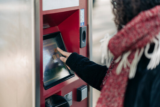 A person with curly hair and a red scarf uses a red ticket or payment machine. Their hand is on the touchscreen, making a selection or completing a transaction.