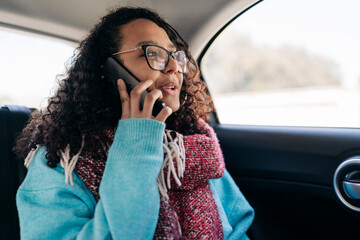 A young woman with curly hair and glasses sits in the back seat of a car, talking on her mobile phone. She wears a blue sweater and a red scarf. This image captures communication on the go.