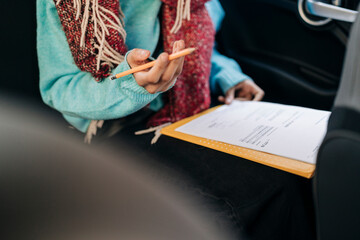 An individual wearing a blue sweater and a colorful scarf holds a pencil, examining papers on a yellow clipboard. They are working on documents inside a vehicle.