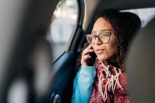 A young woman with curly hair and glasses sits in the back seat of a car, looking out the window while talking on her mobile phone. She wears a blue sweater and a red scarf.