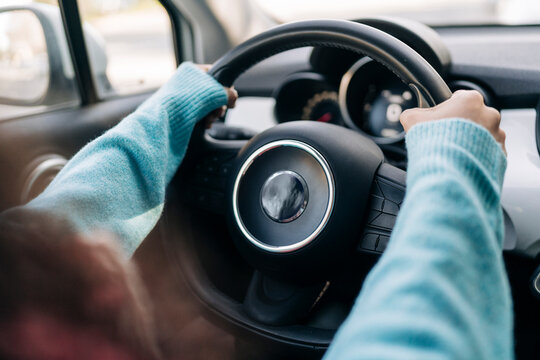 A person wearing a light blue sweater is driving a car. Their hands are firmly on the steering wheel, navigating the vehicle. The dashboard and side mirror are visible, indicating movement.