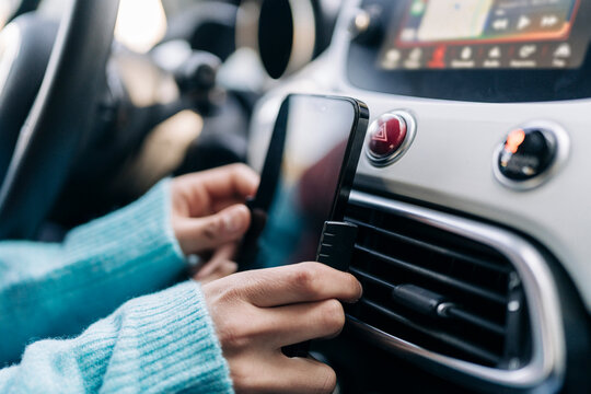 woman wearing a blue sweater is carefully placing a black smartphone into a car mount. The mount is securely attached to the vehicle's air conditioning vent, with the dashboard in the background.