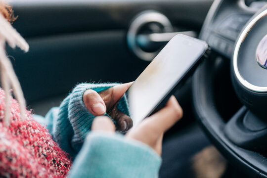 A person wearing a blue knitted sweater and a red top is inside a car. They are holding a smartphone with both hands, actively engaging with the device.