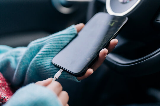 A person in a blue sweater holds a smartphone, plugging in a charging cable. The phone is charging inside a car, with the steering wheel in the background.