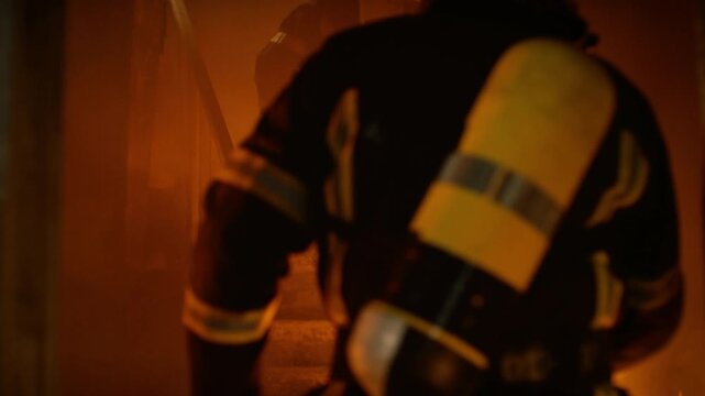 Two Firefighters in Full Protective Gear Run up a Smoky Staircase Inside a Burning Building, Surrounded by Intense Flames and Heavy Smoke, Moving Quickly to Reach People Trapped on the Upper Floor.