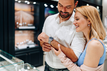 Elegant couple enjoying a moment in a jewelry store, trying on luxury watches while smiling and embracing, making a thoughtful purchase decision for a special occasion