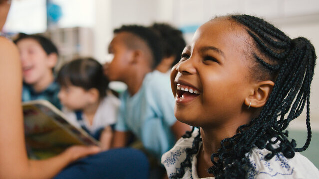 Portrait Of Laughing Female Elementary School Pupil With Teacher Reading To Class In Background