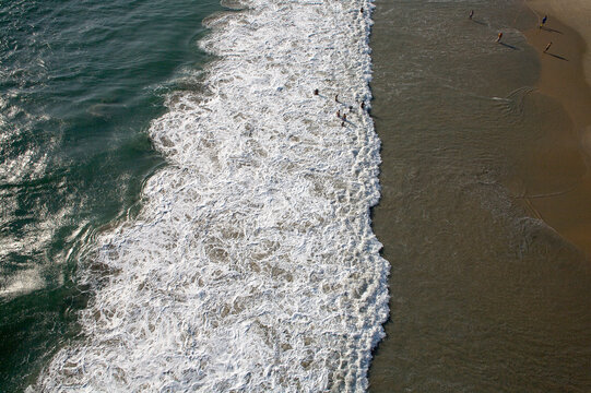 Aerial view of turquoise waters crashing in foamy white waves against the golden sands where tiny figures stroll, Varkala, Kerala, India.