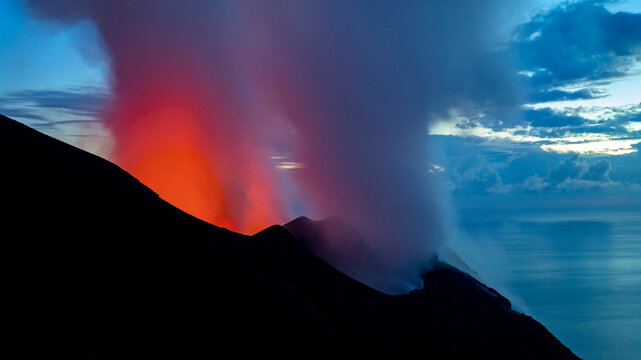 Volcanic eruption at dusk, with bright orange lava and smoke plumes against a blue sky. Stromboli Island, Sicily, Italy