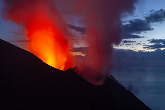 Volcano erupting at night, with bright orange lava and smoke plumes against a dark sky with clouds. Stromboli Island, Sicily, Italy
