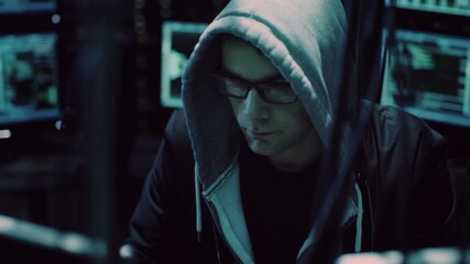 Hacker in a Hoodie and Glasses Sits in a Dark Room, Working on Multiple Monitors Displaying Code, Data Breach, Malware, Spyware. Cyberattack Planning, Identity Theft, and Digital Security Threats - Powered by Adobe