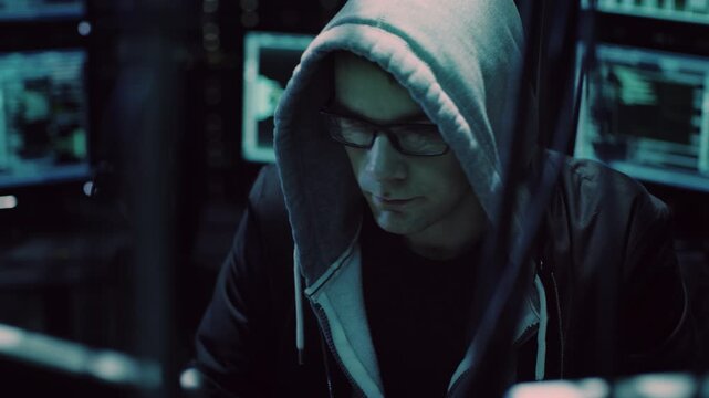 Hacker in a Hoodie and Glasses Sits in a Dark Room, Working on Multiple Monitors Displaying Code, Data Breach, Malware, Spyware. Cyberattack Planning, Identity Theft, and Digital Security Threats