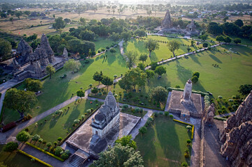 Aerial view of ancient temples standing majestically amidst lush greenery, their carved stones glowing softly in the warm light, Khajuraho, Madhya Pradesh, India.