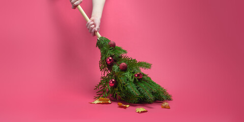 Hands sweep autumn leaves with creative Christmas spruce broom on pink backdrop, hinting at holiday sales, discounts, deals, savings.