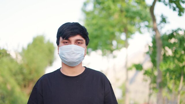 4K Portrait shot of an Indian man wearing Covid 19 surgical mask staring at the camera while standing outdoors. Man stares at the camera while standing in front of mountains during his trip.