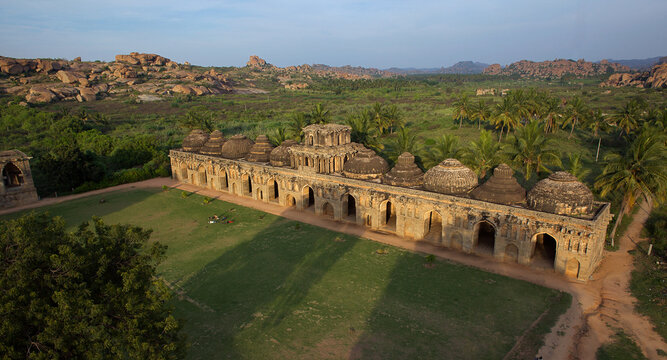 Aerial view of the ancient Elephant Stables standing proudly amidst green fields and rocky terrain under the soft glow of dawn, Hampi, Karnataka, India.