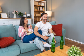 Obraz premium Young couple man and woman sitting at home on sofa taking a break after stressful work day watching sport game or movie on TV, eating popcorn and drinking beer.