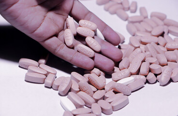 A striking macro shot of a hand scooping up a pile of light-colored, oval-shaped pills or capsules scattered on a white surface.
