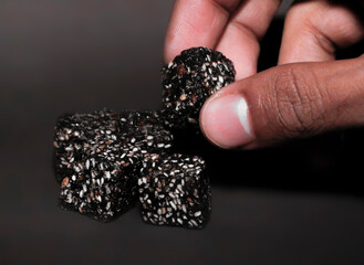 A Detailed Close-up of a Human Hand Delicately Holding a Single, Dark, Rustic Cube, a Healthy and Natural Snack Made from a Rich, Textured Paste and Generously Embedded with White Sesame Seeds