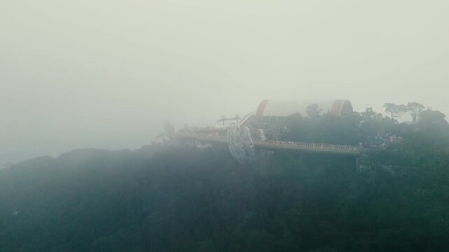 Golden pedestrian bridge spanning misty landscape, supported by massive stone hands at ba na hills landmark near da nang, vietnam, drawing worldwide travelers