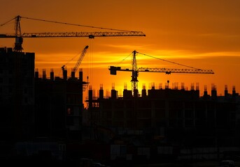 Silhouetted construction cranes stand against a vibrant orange sunset sky