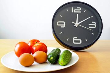 Circadian nutrition timing: tomatoes, cucumbers and eggs arranged on plate next to the clock at noon. Concepts of chrono-diet, intermittent fasting and balanced eating window. Mindful meal planning