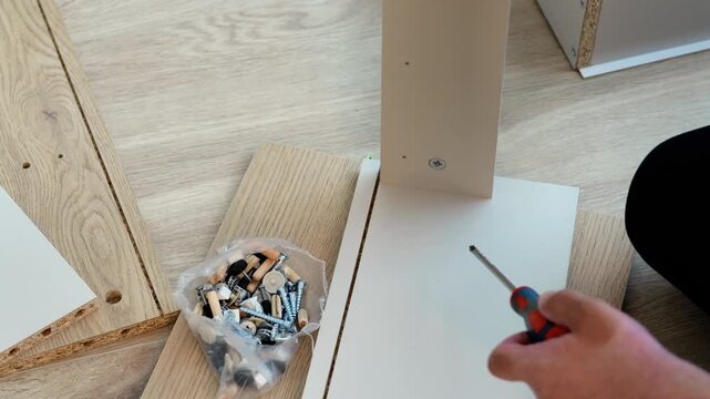 A top-down view of a hand using a screwdriver to tighten a screw into a piece of white flat-pack furniture. A bag of assorted hardware is visible next to the piece.