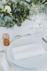 Elegant wedding table setting with white plate, folded napkin, wine glasses, and a small caramel jar as gift.