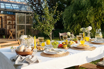 Outdoor table set for meal featuring plates with food, glasses of orange juice, bread, fruit, and flowers arranged on tablecloth in garden near glass greenhouse