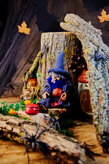 Fototapeta premium a perky halloween witch in a pointed hat with spiders, bottles, a saucepan and a ladle sits on a stump under a snag to brew a witch's potion in a gloomy forest on All Saints Day, selective focus