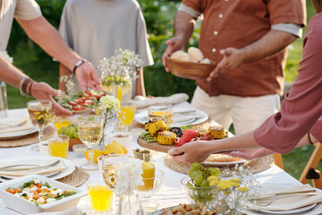Multiethnic group of men and women serving grilled vegetables, salad, bread and drinks at outdoor table, hands reaching for food, enjoying summer gathering
