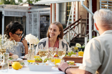 Caucasian young woman smiling while sitting at outdoor table with boy and senior Caucasian man, sharing meal and engaging in conversation during family gathering