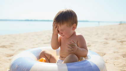 Baby boy sitting in inflatable ring crying with hand on face. Concept of discomfort, emotions,...
