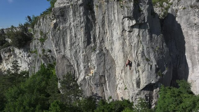 AERIAL: Wide perspective of a male climber high up on a sheer rock wall above lush green treetops. Climbing area full of diverse climbing routes and scenic views in the middle of the karst landscape.
