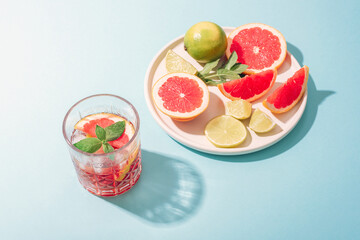 Glass of grapefruit drink with lime slices and mint leaves and plate with fruits on blue background in sunlight. Top view