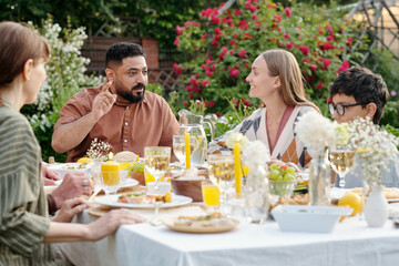 Multiethnic family sitting outdoors sharing meal and conversation at garden table surrounded by flowers and food