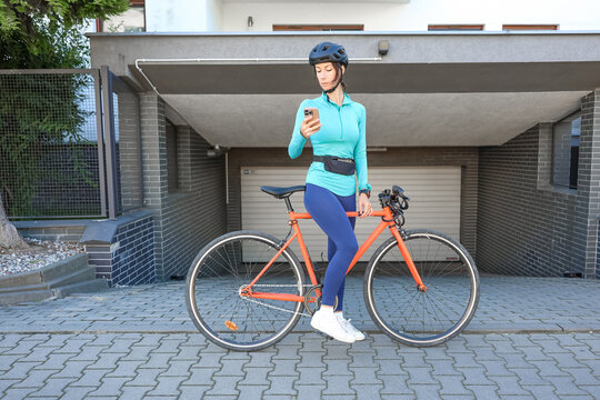 Sporty woman with bicycle in front of garage, wearing helmet and looking at smartphone, setting fitness tracker before cycling ride